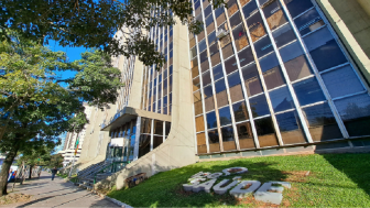 Fachada do prédio do IPE SAÚDE. Com concreto e vidro, na grama, com as letras feitas em placas de cimento, a inscrição "ipe saúde" é visível na diagonal.