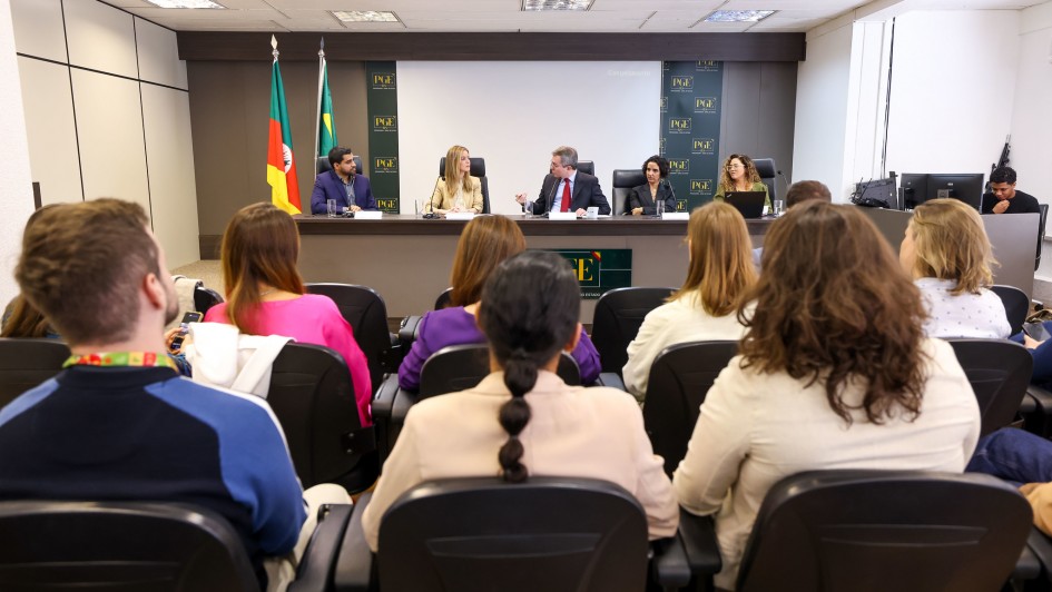 um audit&oacute;rio com participantes sentados, de costas para a foto, assistindo a palestra. No fundo, cinco pessoas, tr&ecirc;s mulheres e dois homens, est&atilde;o sentadas &agrave; mesa, participando da palestra. 