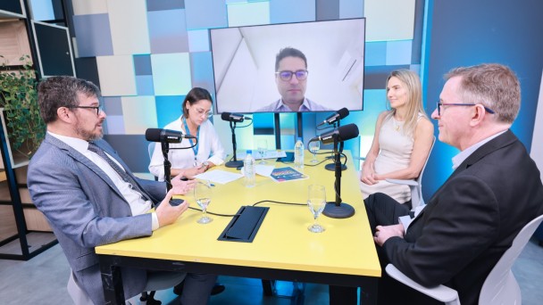 quatro pessoas sentadas &agrave; mesa, em um espa&ccedil;o retangular com decora&ccedil;&atilde;o em tons de azul e branco. No fundo uma TV exibe a imagem de uma quinta pessoa, que participa de forma virtual. 